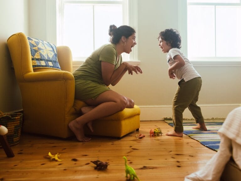 Mother and young child playfully pretending to be dinosaurs together in a bright living room with toy dinosaurs scattered on the floor.