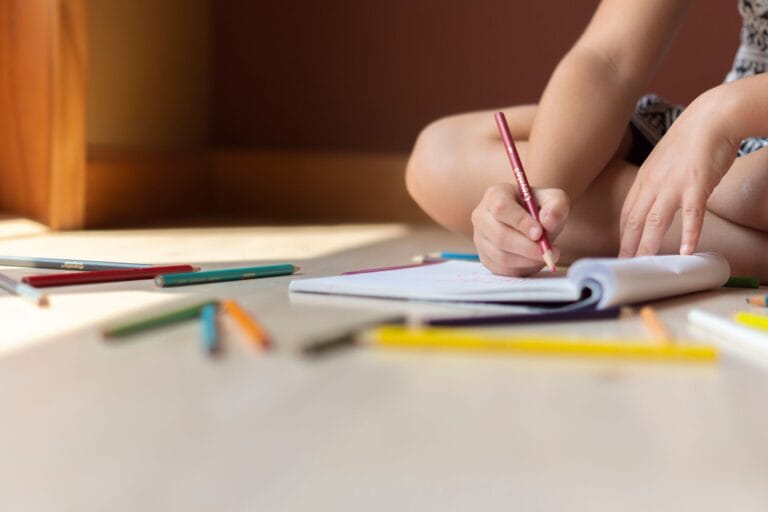 Crop anonymous child in casual clothes sitting on floor with legs crossed and drawing in copybook by using multicolored pencils