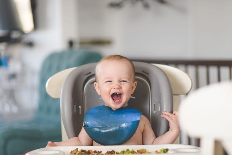 Happy baby laughing in a high chair during mealtime, wearing a blue bib.